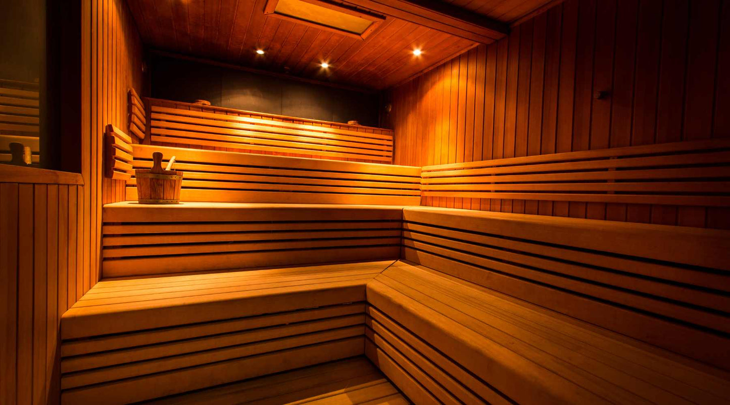 The image depicts the interior of a sauna with wooden benches and walls. There's a gentle glow from the lights, illuminating the warm tones of the wood. A traditional sauna bucket and ladle are placed on the lower bench, suggesting readiness for a steam s