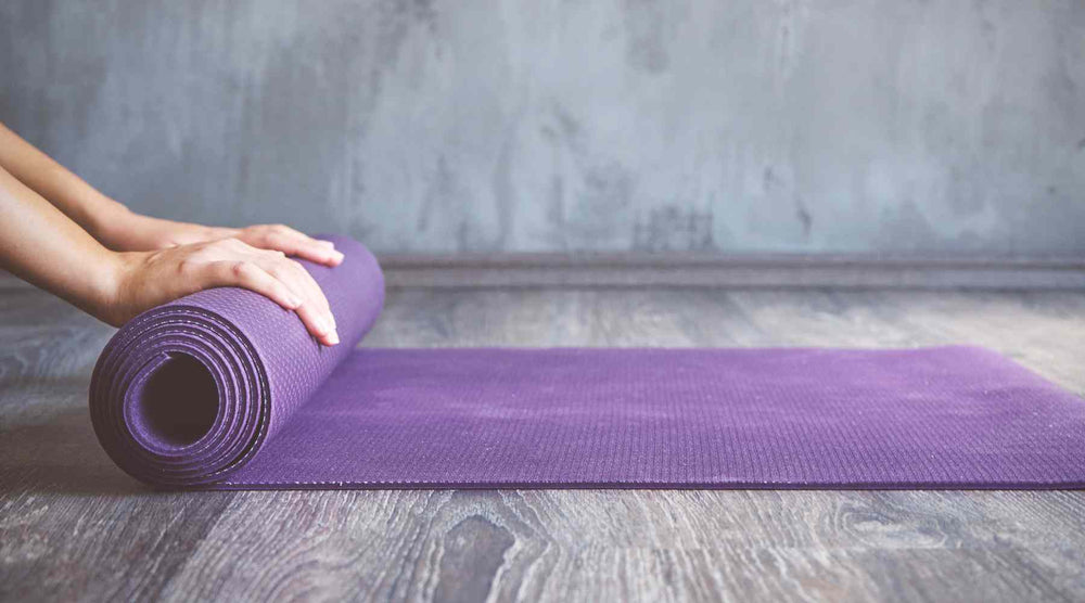 The image shows a rolled-up purple yoga mat being placed on a wooden floor by a person whose hands are visible in the frame. The background features a textured grey wall, creating a calm and minimalist setting that is often associated with yoga or meditat