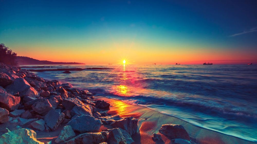 A sunrise over a rocky beach with reflections on the water and ships on the horizon.