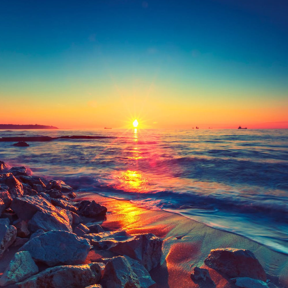 A sunrise over a rocky beach with reflections on the water and ships on the horizon.
