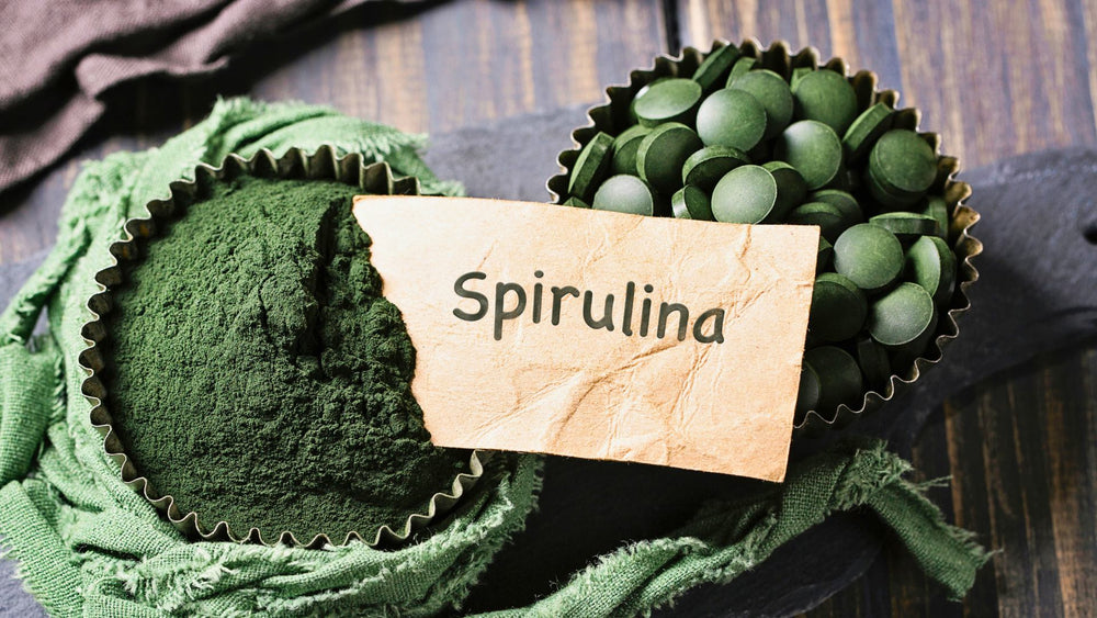 The image shows two containers, one with Spirulina powder and the other with Spirulina tablets, placed on a rustic wooden surface. A torn paper label with the word "Spirulina" 