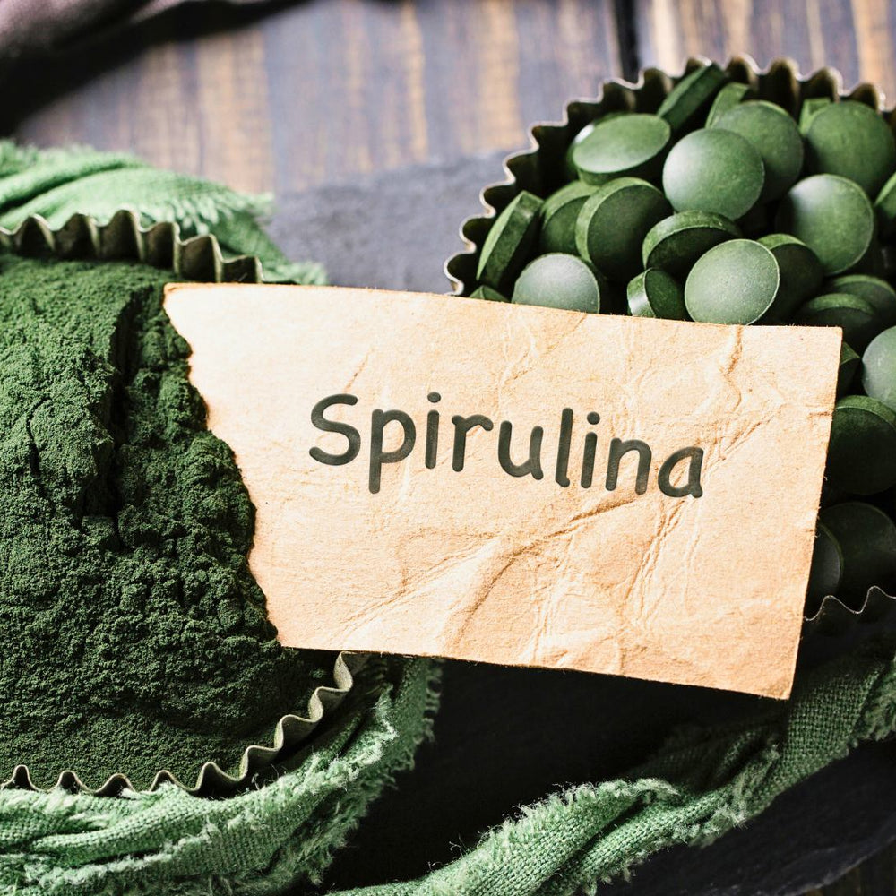 The image shows two containers, one with Spirulina powder and the other with Spirulina tablets, placed on a rustic wooden surface. A torn paper label with the word "Spirulina" 